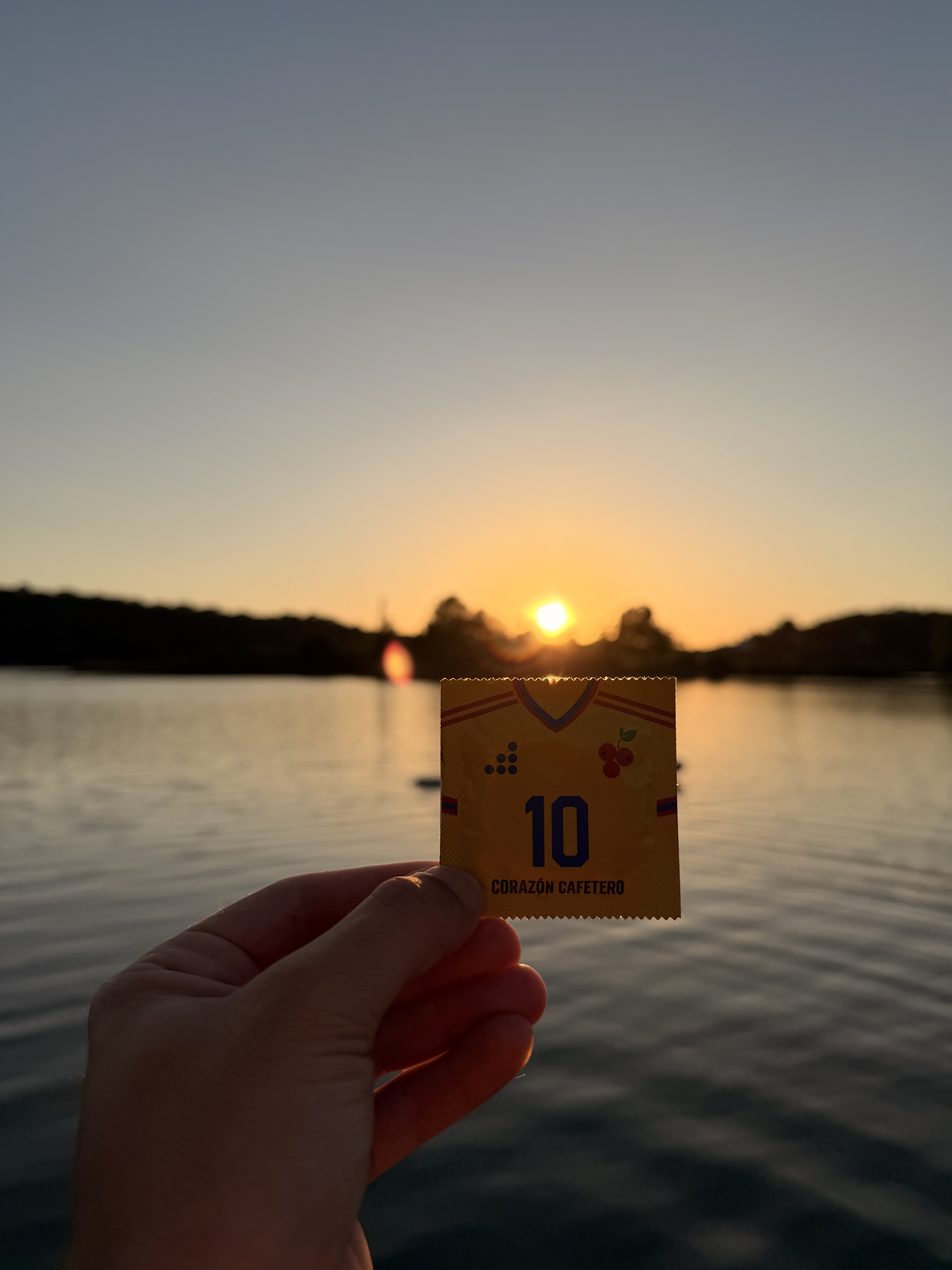Mano sosteniendo el preservativo PreservatiGold Colombia 'Corazón Cafetero nº10' en amarillo al atardecer dorado sobre el agua