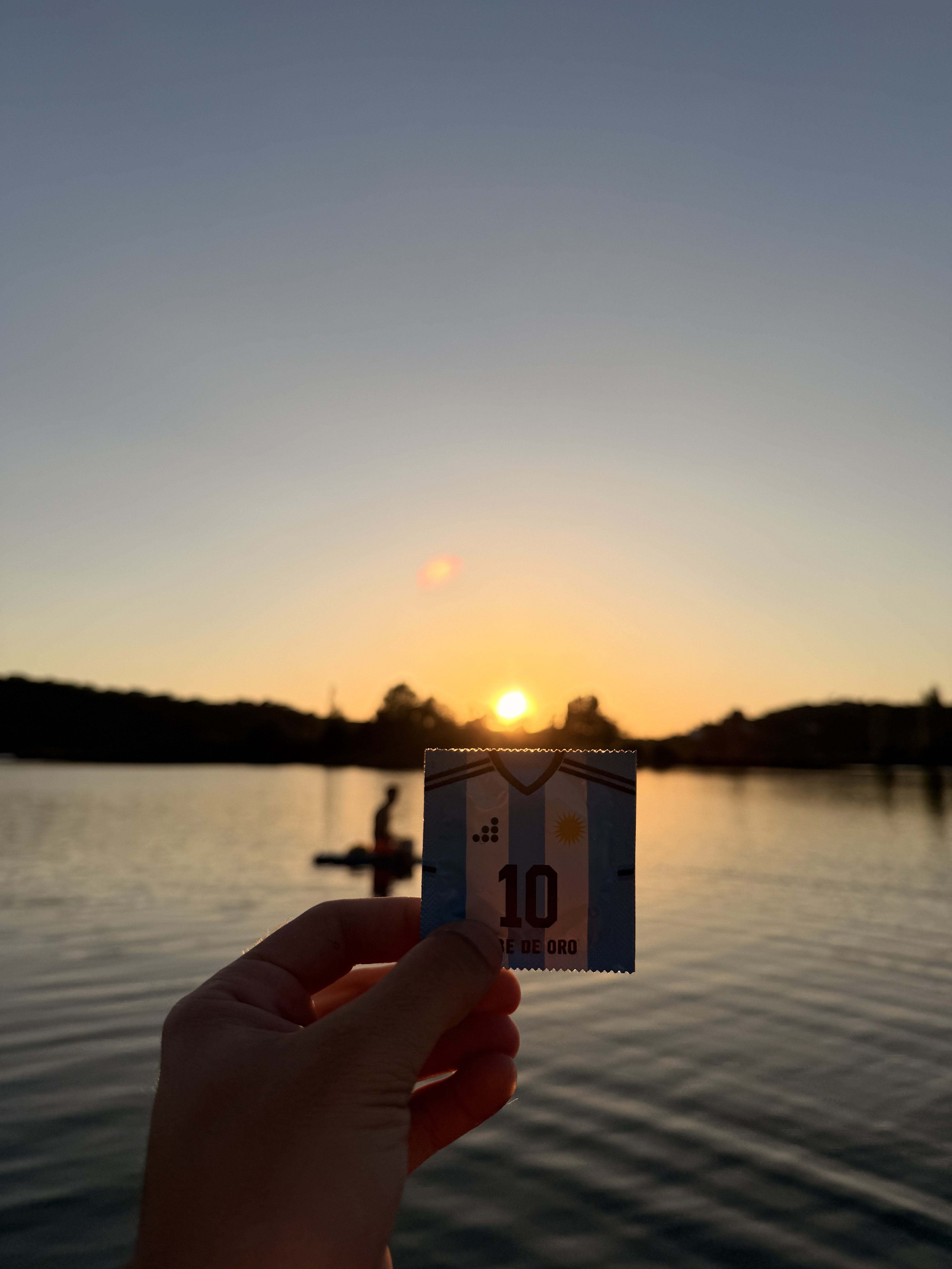 Mano sosteniendo el preservativo PreservatiGold Argentina 'Pibe de Oro nº10' con los colores albicelestes al atardecer sobre un lago